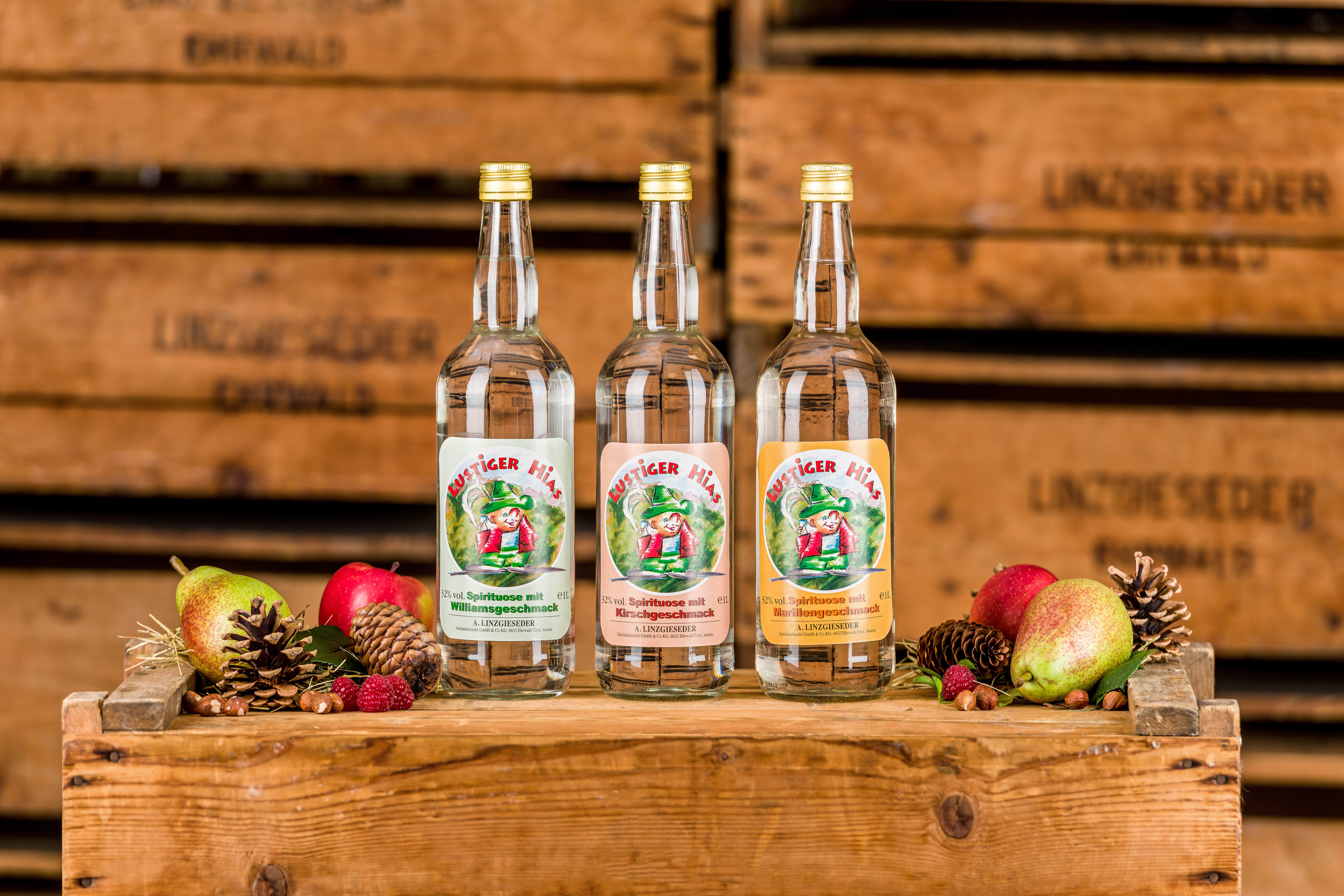 Drei Flaschen der Spirituose Lustiger Hias stehen auf einer Holzkiste. Links und rechts daneben liegen Tannenzapfen, Äpfel, Birnen und Nüsse. Im Hintergrund sind Holzkisten mit dem Linzgieseder Brandzeichen sichtbar, die dem Bild eine rustikale Atmosphäre