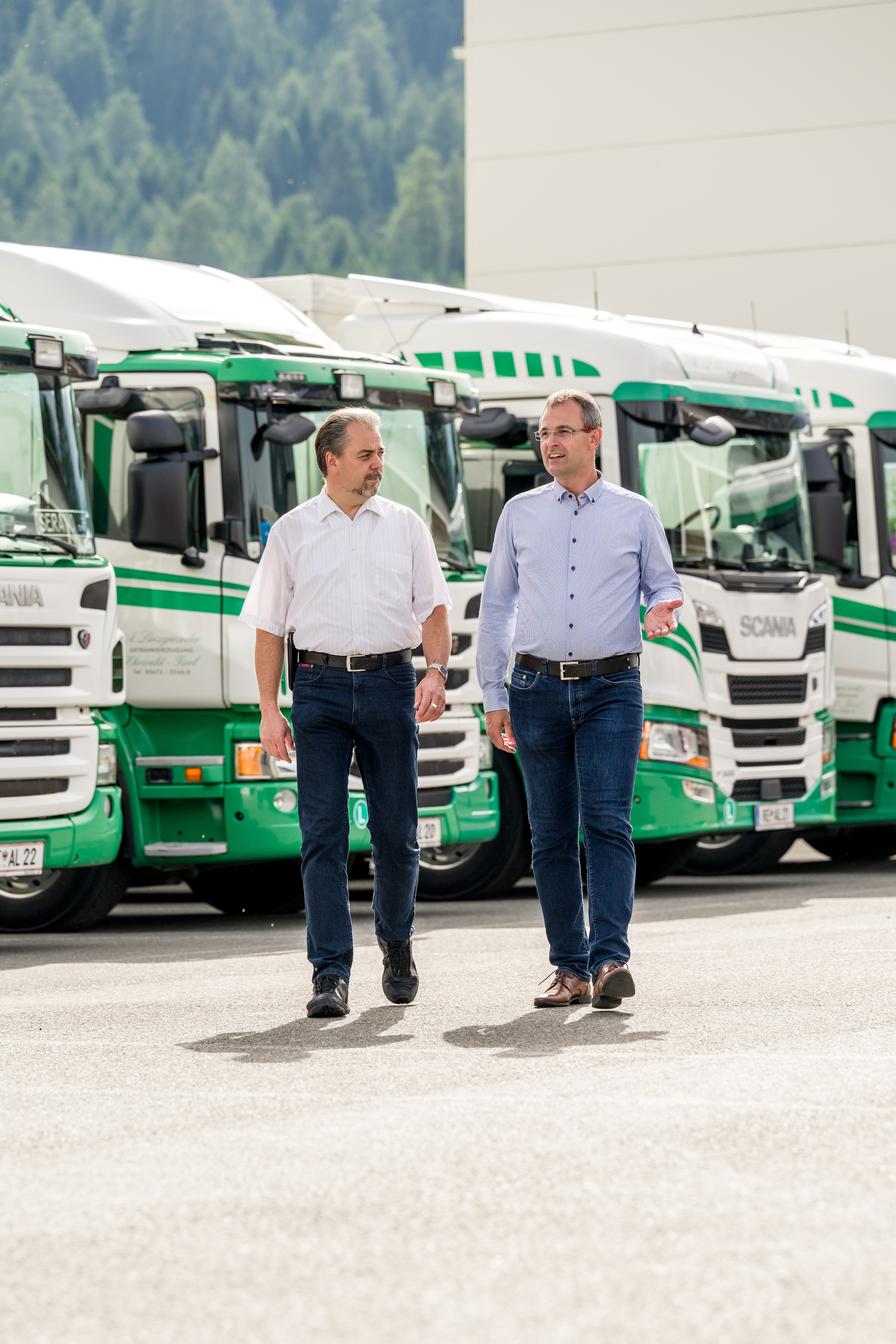 Arthur und Alfred Linzgieseder gehen über das Betriebsgelände. Im Hintergrund parken Lkw der Firma, die ordentlich auf dem Gelände abgestellt sind. Die beiden Männer sind in Bewegung, während die Lkw ruhig im Hintergrund stehen.