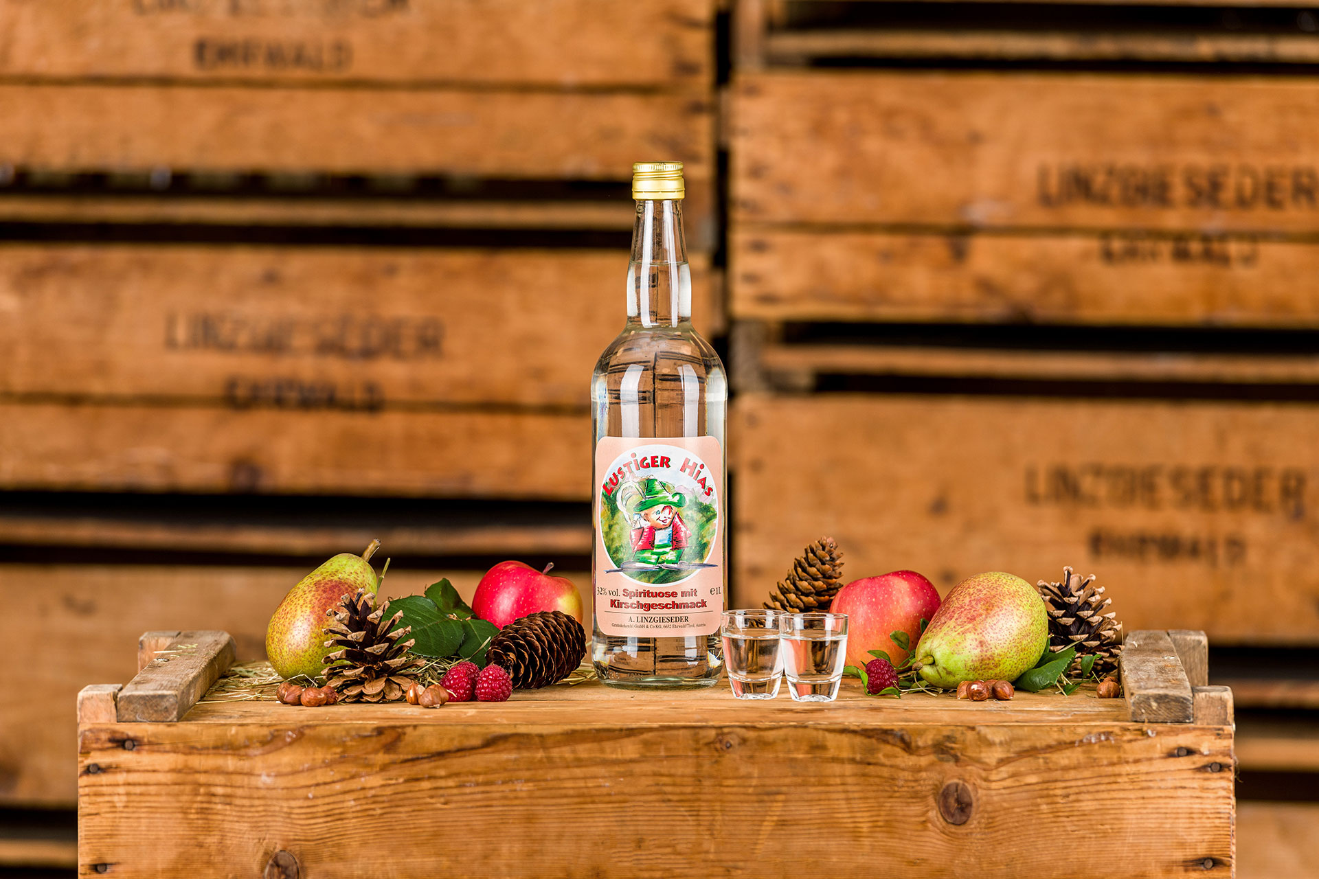 Spirituosenflasche ‚Lustiger Hias Kirsche‘ mit zwei gefüllten Schnapsgläsern mittig auf einer Holzkiste stehend. Links und rechts davon liegen Birnen, Äpfel, Zapfen und Nüsse. Der Hintergrund zeigt gestapelte Holzkisten mit Linzgieseder-Aufschrift.
