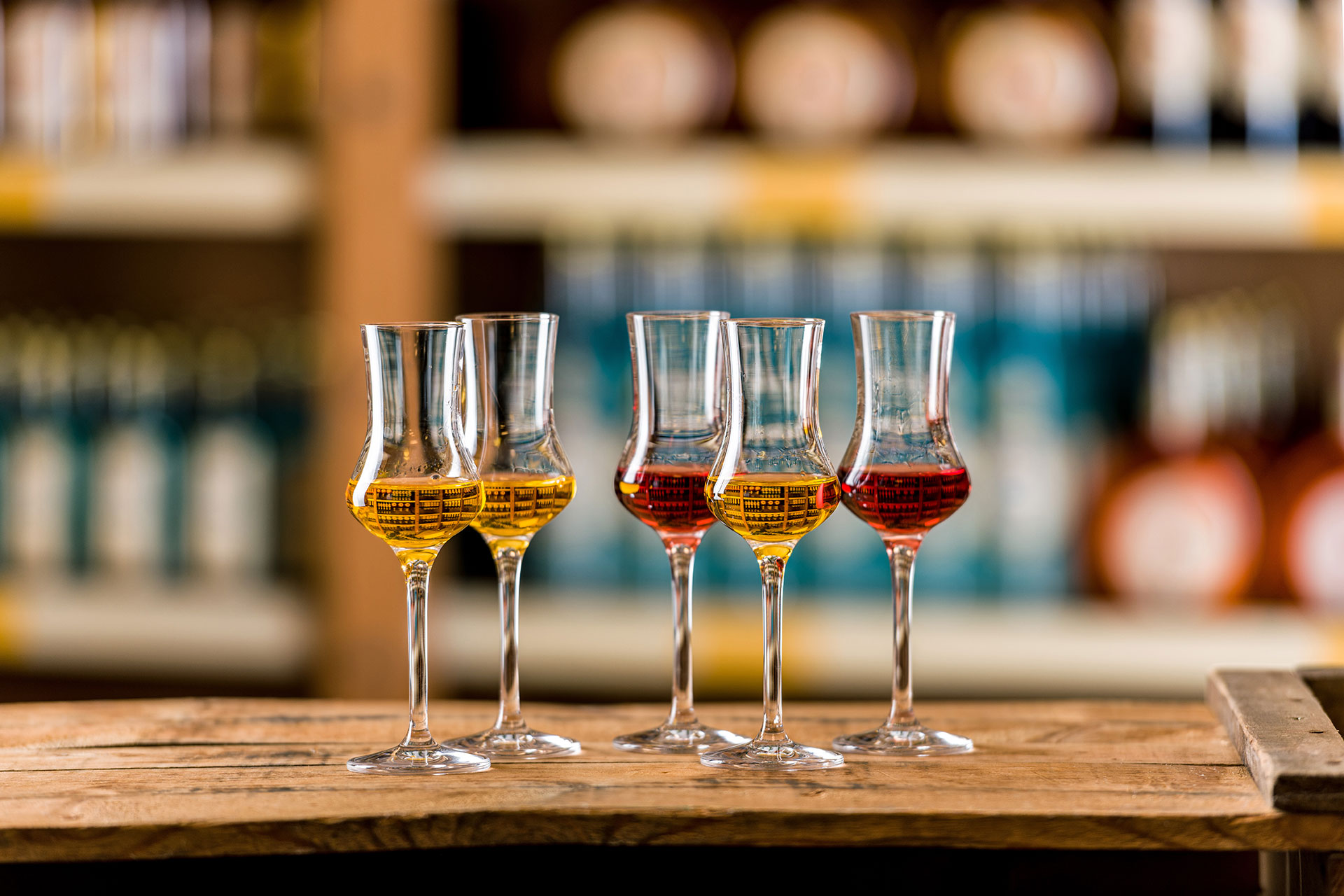 Fünf gefüllte, goldgelbe bis dunkelrote Selektion-Schnapsgläser stehen auf einer Holzkiste. Der Hintergrund zeigt verschwommene Regale mit Spirituosen.