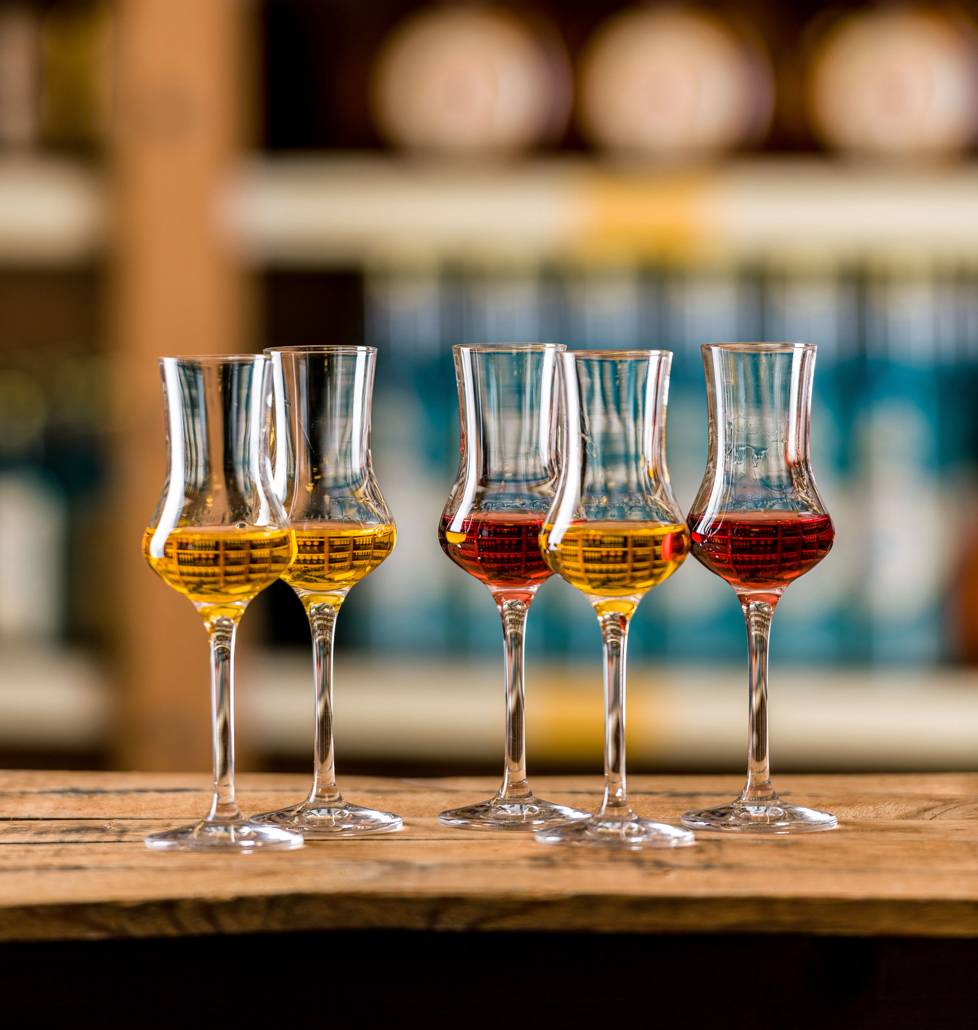 Fünf gefüllte, goldgelbe bis dunkelrote Selektion-Schnapsgläser stehen auf einer Holzkiste. Der Hintergrund zeigt verschwommene Regale mit Spirituosen.