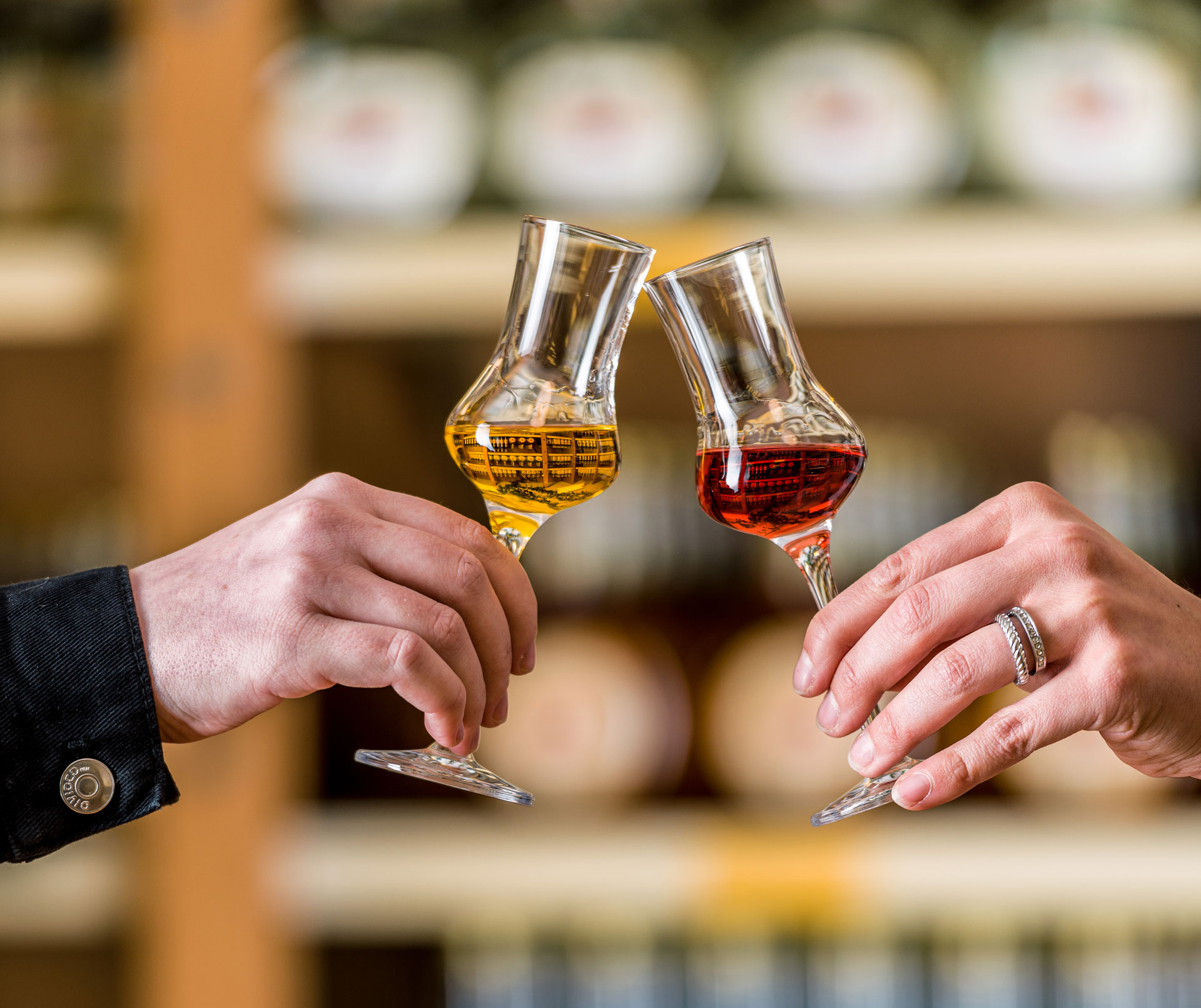 Links hält eine Hand ein Selektionsglas mit goldgelber Flüssigkeit, während in der Mitte des Bildes eine weitere Hand ein zweites Selektionsglas hält und damit anstößt. Das rechte Glas enthält eine rötlich-dunkle Flüssigkeit. Im Hintergrund ist ein Regal 