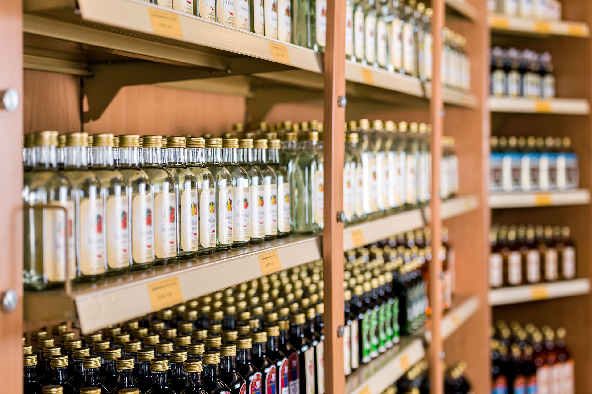 Ein Bild von mehreren Linzgieseder Schnapsflaschen, die ordentlich in einem Regal angeordnet sind. Die Flaschen sind so platziert, dass die Etiketten seitlich sichtbar sind. Das Licht reflektiert auf den Glasflaschen und hebt die Details der Etiketten her