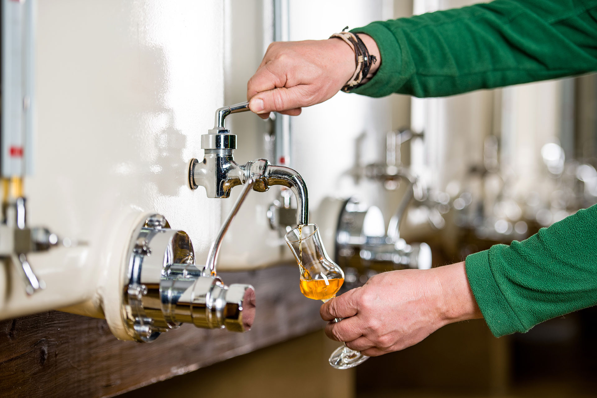Fokus auf den Abfüllhahn eines Stahltanks, der mit Spirituosen befüllt ist. Zwei Hände bedienen den Abfüllhahn, aus dem eine gelbliche Flüssigkeit in ein Selektionsglas rinnt und im Glas leuchtet.