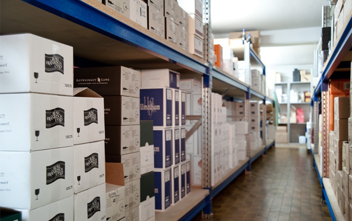 Blick ins Weinlager durch einen Gang zwischen blauen Schwerlastregalen. Der Fokus liegt auf dem linken Regal, das voll mit verschiedensten, fein sortierten Kartons in und auf den Regalen ist.