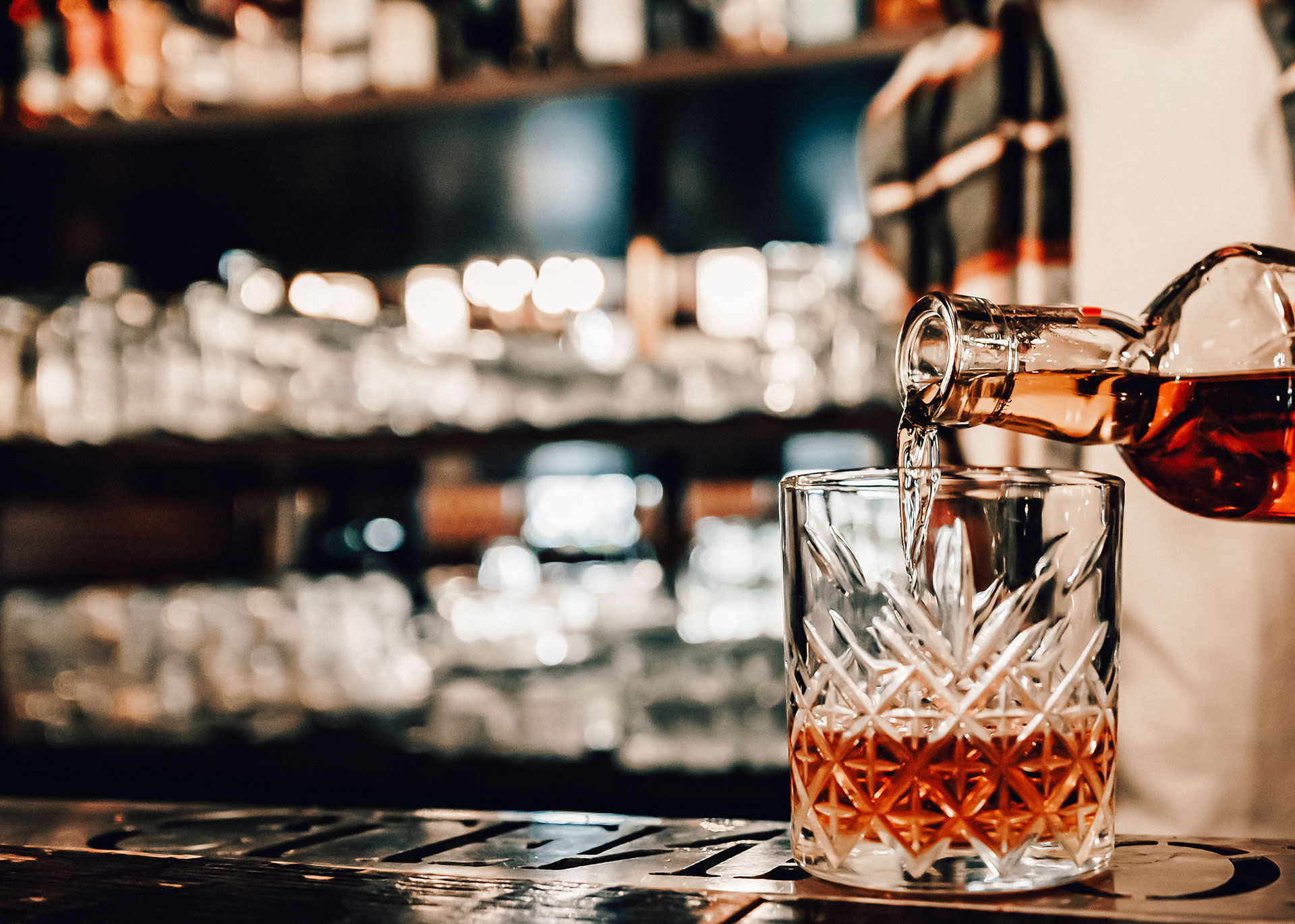 Im Bild, rechts im Fokus, ein Whiskey-Tumbler, der gerade von einer ins Bild ragenden Flasche mit einer gold-rot schimmernden Flüssigkeit befüllt wird. Der Hintergrund ist unscharf, wobei die Umrisse einer Bar zu erkennen sind