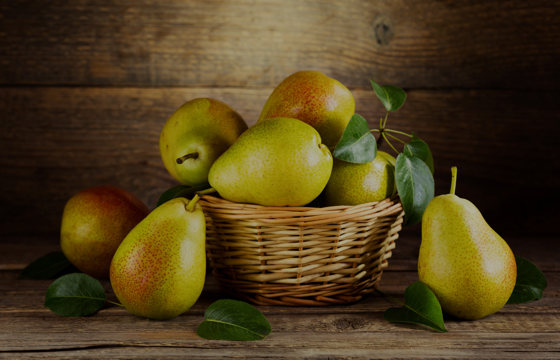 Frontansicht eines Korbs mit Birnen, dekorativ umgeben von Birnenblättern. Links neben dem Korb liegen zwei Birnen auf einer Holzoberfläche, rechts eine Birne. Der Hintergrund besteht aus Holzdielen.