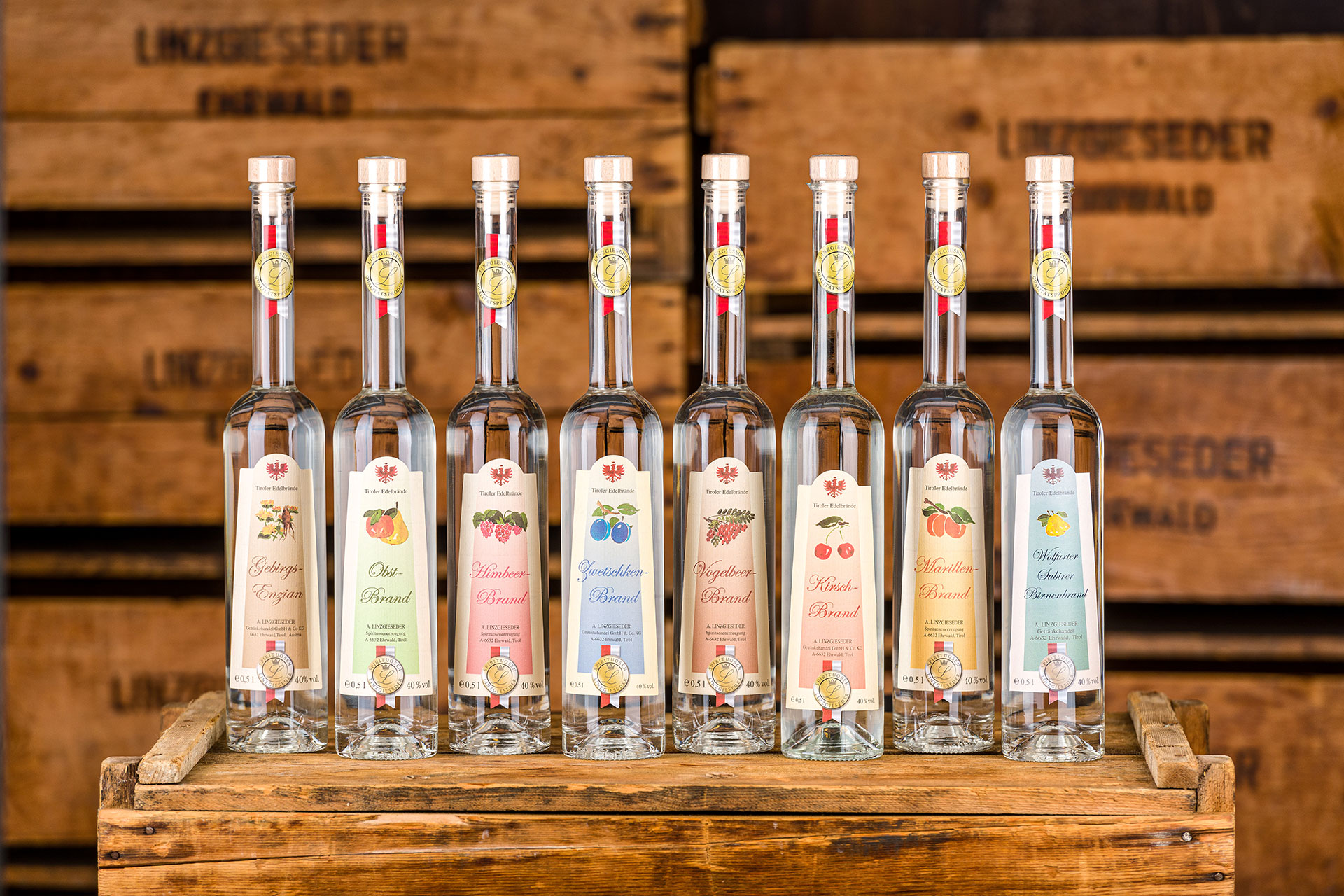 Acht langhälsige Flaschen der Linzgieseder Edelbrände stehen auf einer Holzkiste, im Hintergrund sind weitere Holzkisten mit Linzgieseder Branding zu sehen.