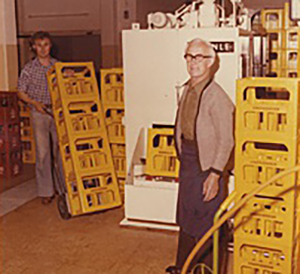 Ein Farbbild, das ein Lager mit gelben Kisten zeigt. Zwei Personen blicken direkt in die Kamera. Die Szene vermittelt eine alte Atmosphäre, die dem Bild einen historischen Charme verleiht