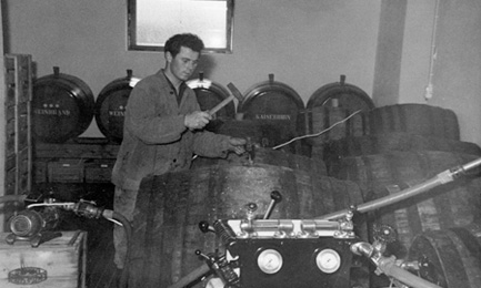 Ein schwarz-weißes Bild von Alfred Linzgieseder Senior, der ein Fass mit einem Hammer verschließt. Einige Fässer sind im Hintergrund und Vordergrund des Bildes zu sehen, was eine arbeitsreiche Atmosphäre schafft.