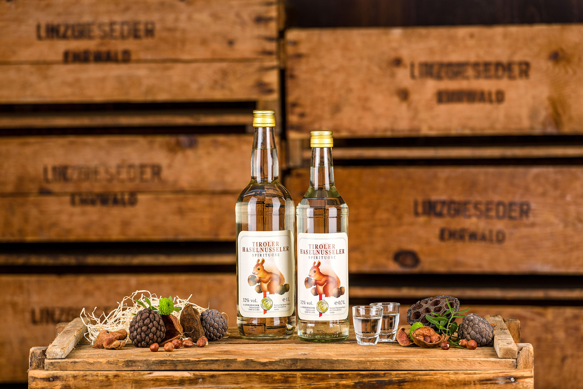 Zwei Spirituosenflaschen ‚Tiroler Haselnusseler‘, eine 1l und eine 0,75l, mit zwei gefüllten Schnapsgläsern mittig auf einer Holzkiste stehend. Links und rechts davon liegen Zapfen und Nüsse. Der Hintergrund zeigt gestapelte Holzkisten mit Linzgieseder-Au