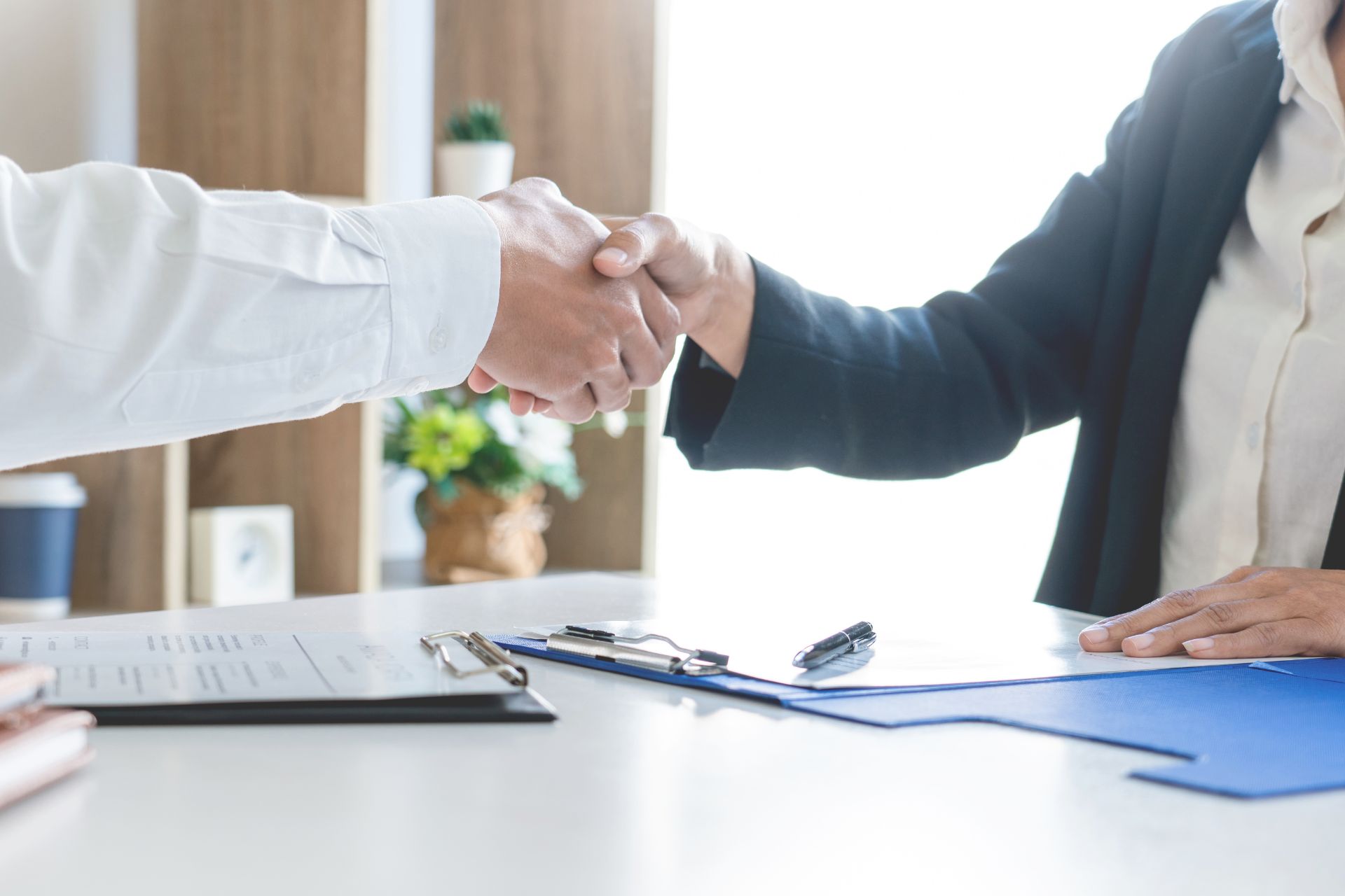 Zwei formell gekleidete Personen geben sich die Hand in einem professionellen Bürosetting. Auf dem Tisch liegt eine Bewerbungsmappe sowie ein kleiner Flipchart. Der unscharfe Hintergrund deutet auf eine geschäftliche Umgebung hin.