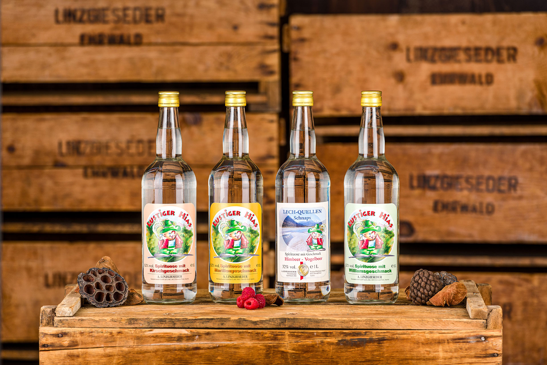 Vier Flaschen der Spirituose Lustiger Hias stehen auf einer Holzkiste, von links nach rechts: 1l Flasche mit Kirschgeschmack, 1l Flasche mit Marillengeschmack, 1l Flasche Lech-Quellen Schnaps und 1l Flasche mit Williamsgeschmack. Links und rechts daneben 