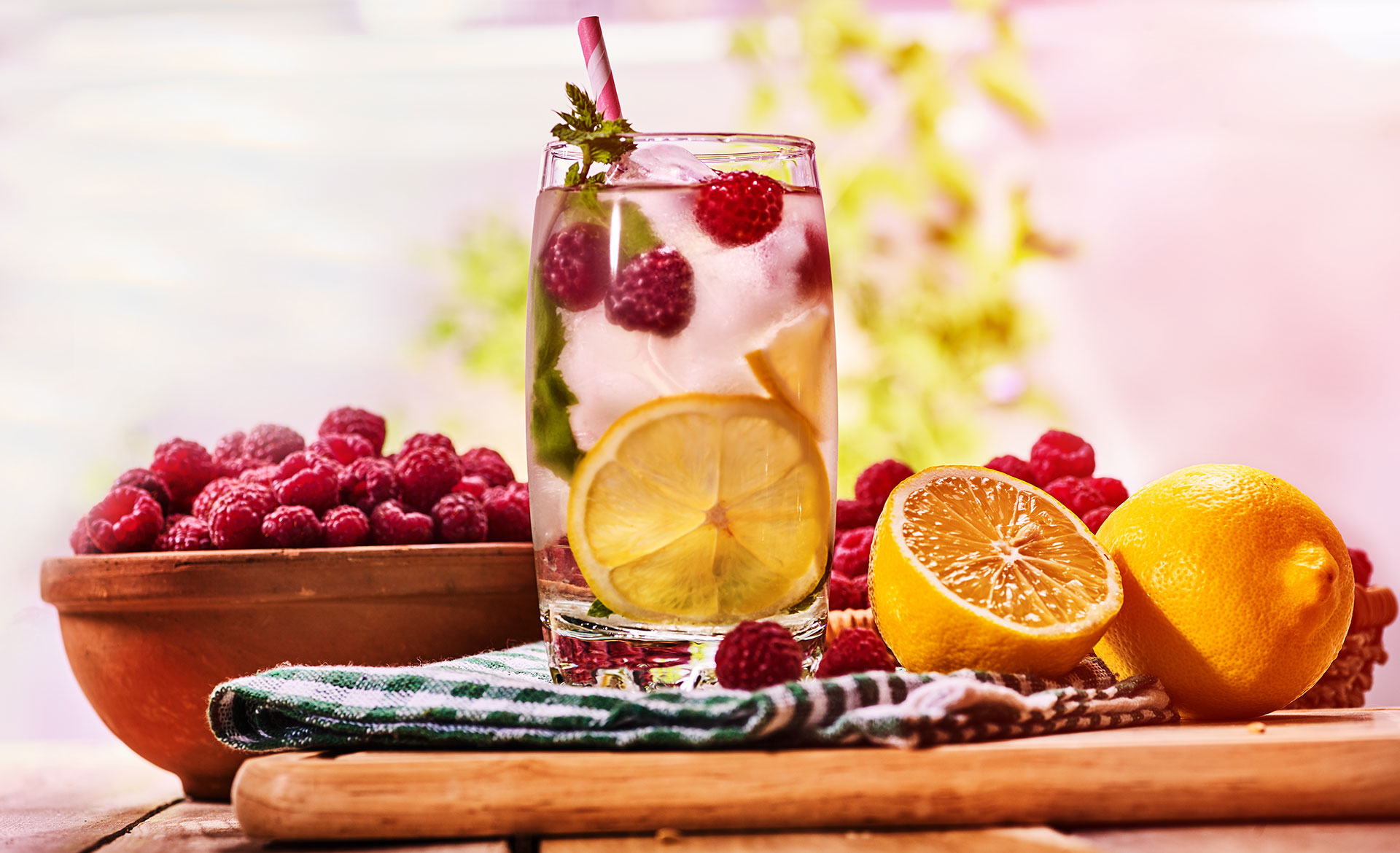 Querformat-Bild, im linken Drittel eine Holzschale, gefüllt mit Himbeeren, leicht verdeckt von einem Jausenbrett, auf dem ein Geschirrtuch liegt. Darauf steht ein Wasserglas, gefüllt mit Eiswürfeln, Himbeeren und Zitronenscheiben. Rechts daneben liegt ein