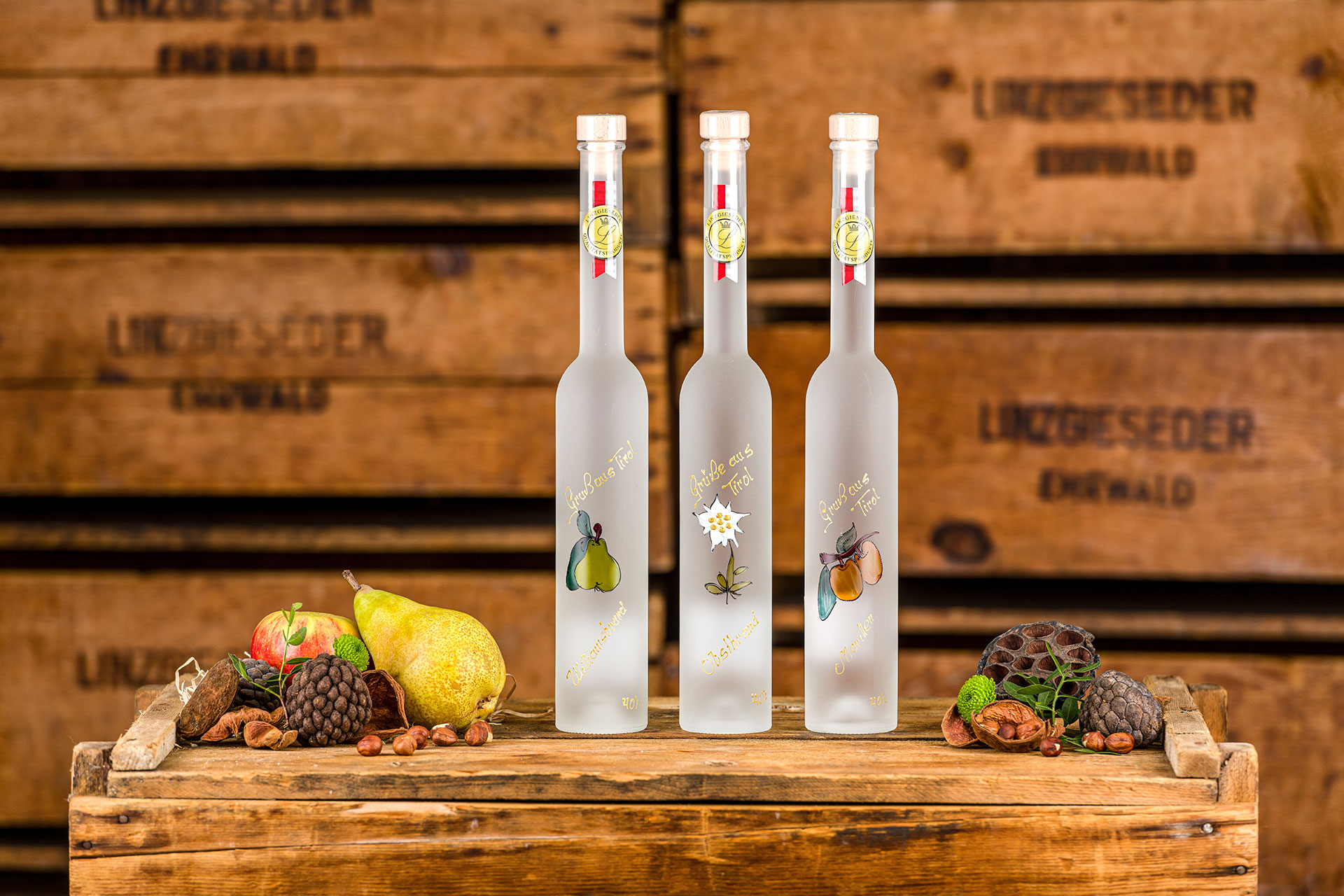 Drei hohe, schmale Flaschen in mattem Weiß mit Windowcolor-Bildern: links eine Birne, mittig ein Edelweiss und rechts Marillen. Die Flaschen stehen auf einer Kiste, daneben sind Obst, Nüsse und Zapfen zur Dekoration arrangiert. Der Hintergrund wird von ge