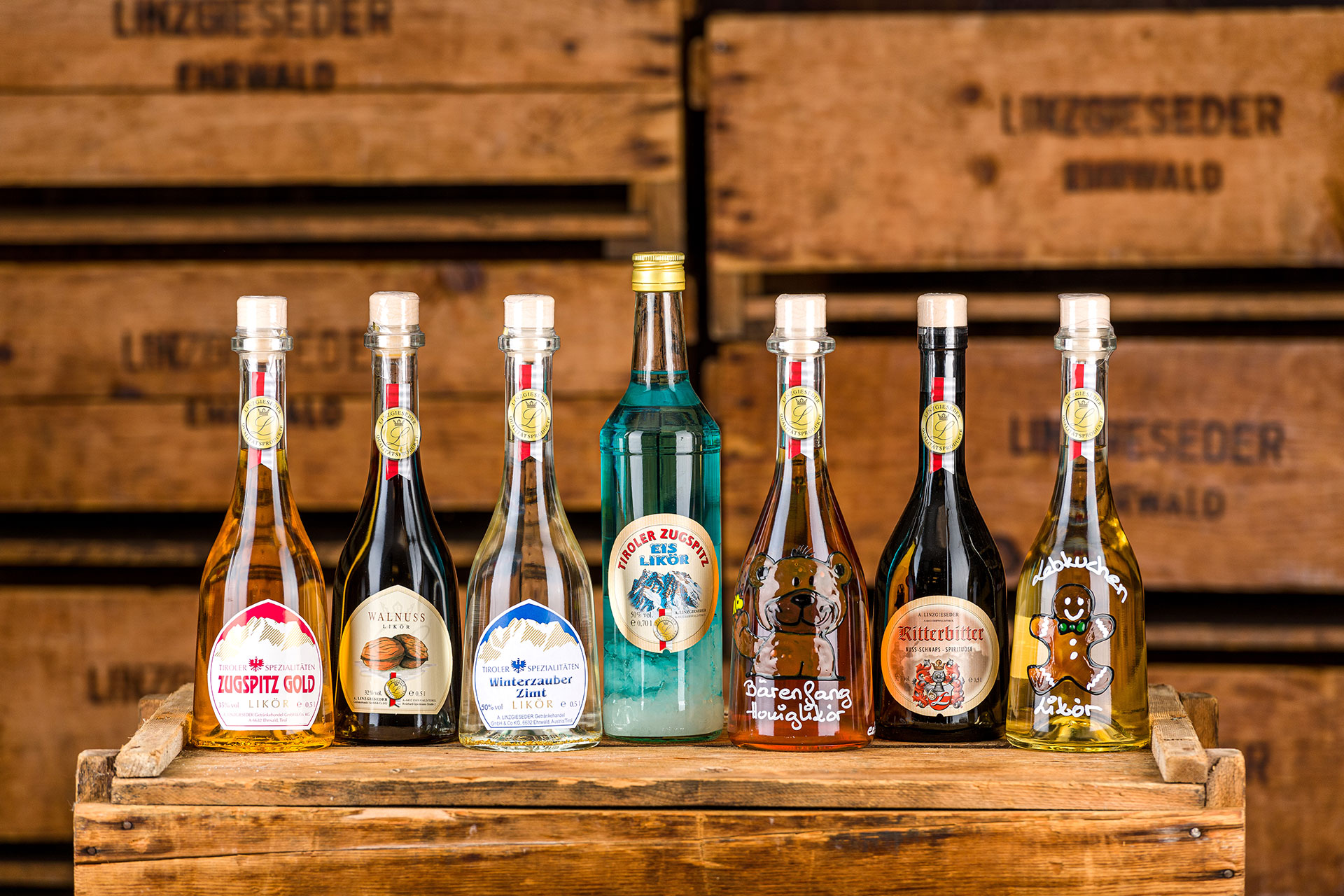 Sieben verschiedene Spirituosenflaschen von links nach rechts: Zugspitzgoldlikör, Walnusskräuterlikör, Winterzauberlikör, Zugspitz Eislikör, Bärenfang Likör, Ritterbitterlikör und Lebkuchenlikör, stehend auf einer Holzkiste. Der Hintergrund zeigt Holzkist