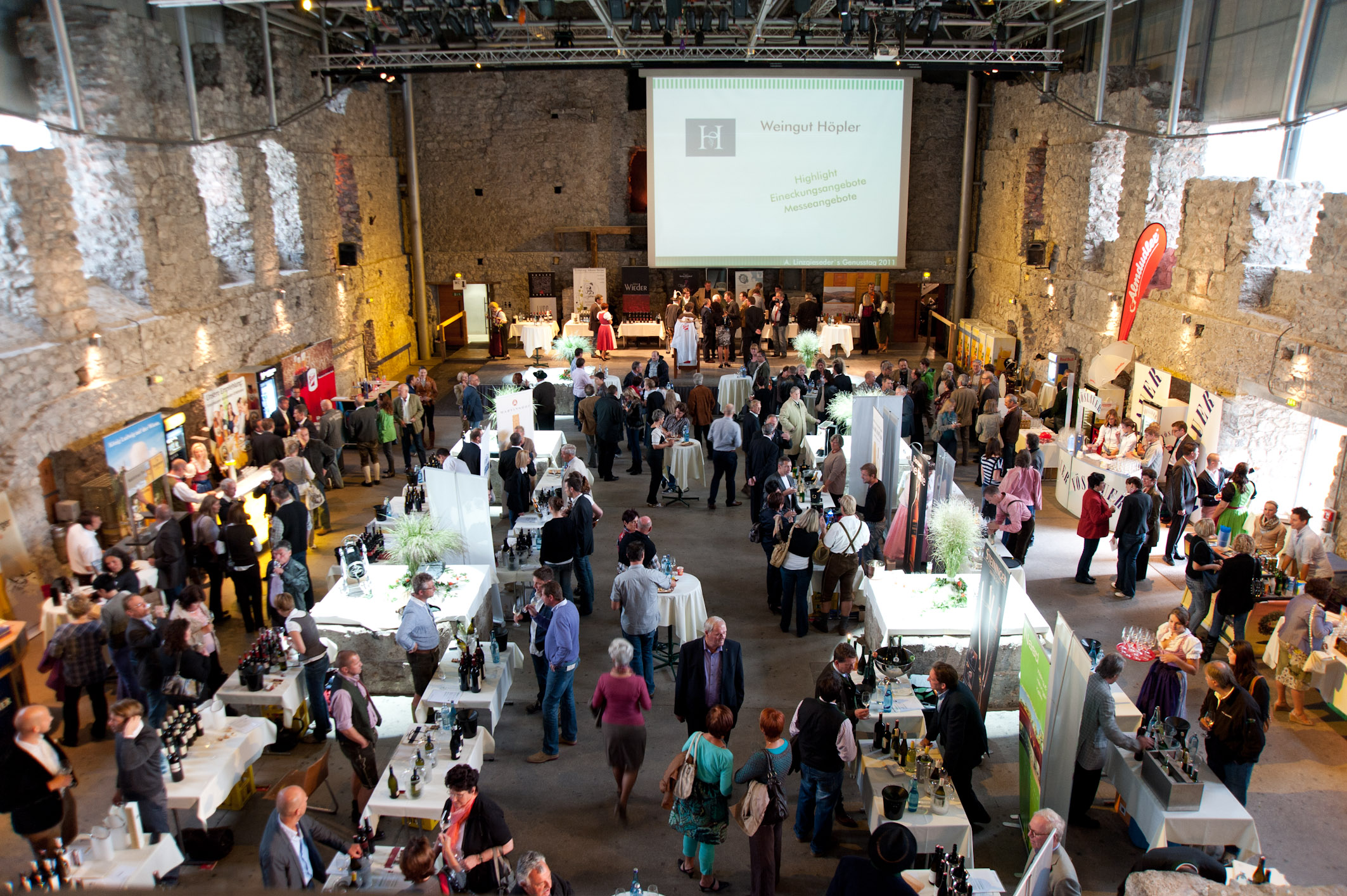 Blick von oben in eine Halle mit vielen Menschen und aufgebauten Ständen. Im Hintergrund ist eine Bühne mit einer großen Leinwand zu sehen.